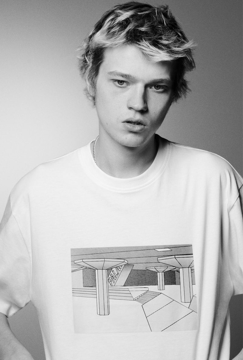 Black and white studio shot of a young adult male model wearing a white t-shirt with a black and white architectural image printed on it.