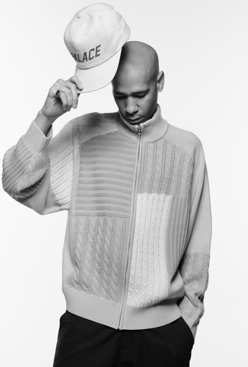 Studio portrait of a bald adult male holding a baseball cap with the word PALACE on it, wearing a patterned zip-up sweater.