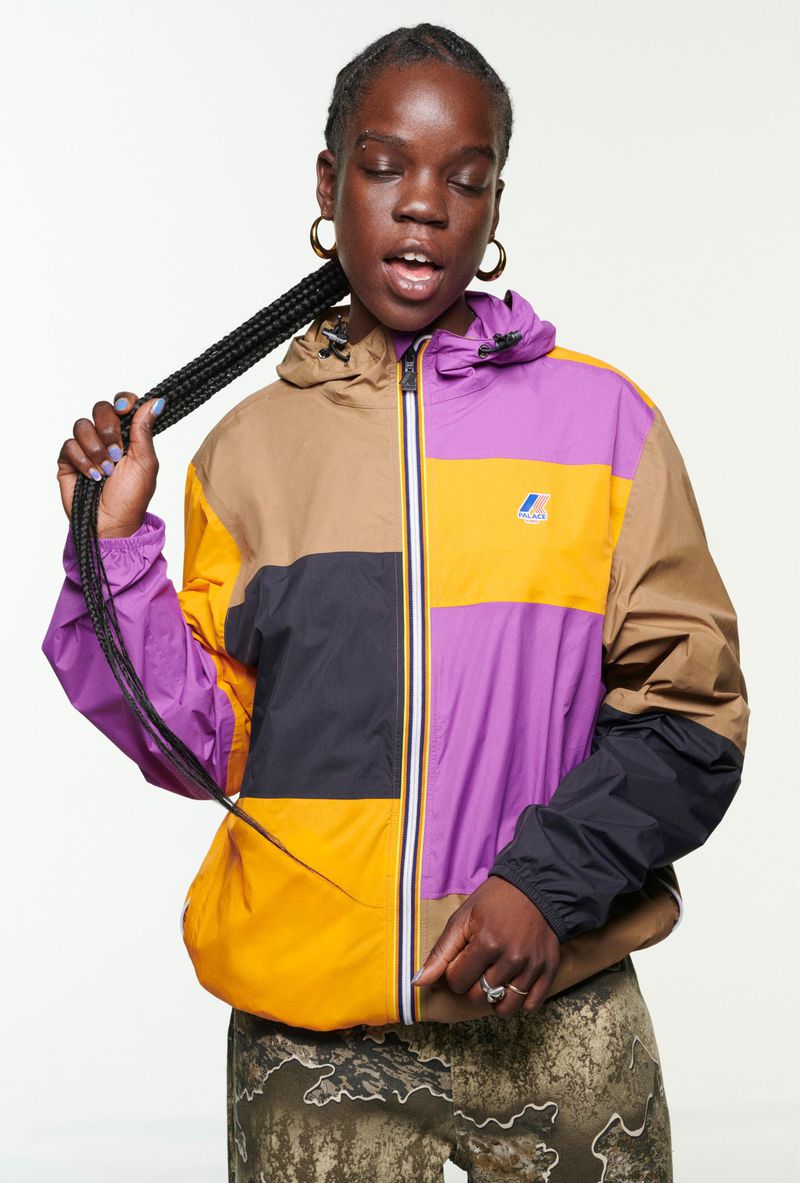A young adult woman poses in a studio wearing a color-blocked windbreaker jacket with a hood and printed pants against a white backdrop.