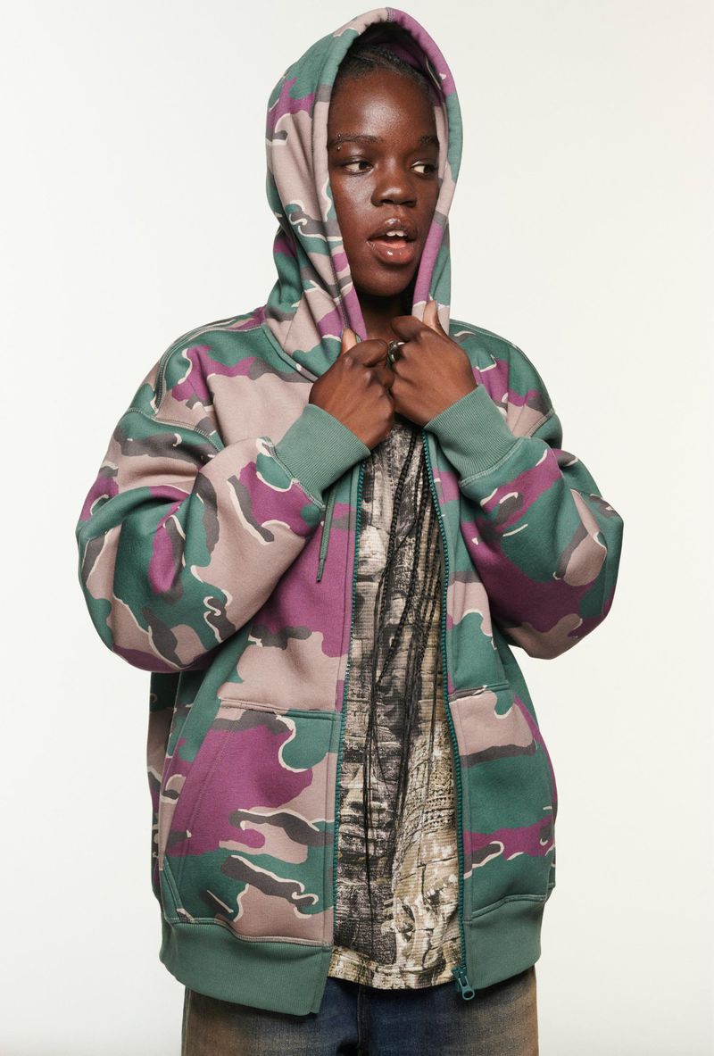 Teen in a camouflage zip-up hoodie with the hood up, standing indoors against a white backdrop, modeling casual fashion.