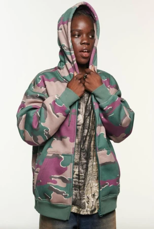 Teen in a camouflage zip-up hoodie with the hood up, standing indoors against a white backdrop, modeling casual fashion.