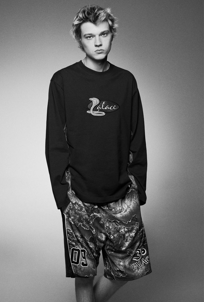 Monochrome portrait of a young adult man wearing a Palace long sleeve shirt and patterned shorts against a light grey backdrop.
