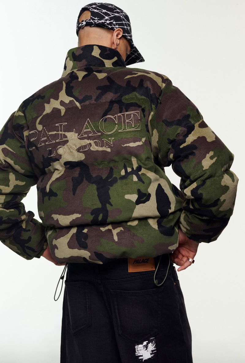 Camo puffer jacket featuring the 'Palace London' logo, worn with dark denim jeans and a black and white baseball cap in a studio setting.