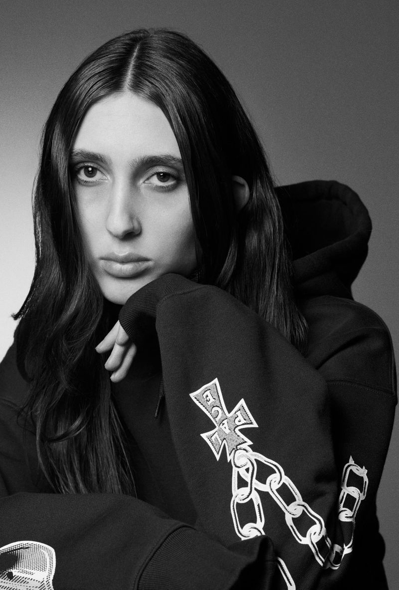 Portrait of a young adult female with long, dark hair wearing a black hooded sweatshirt with graphic elements, set against a grey background.