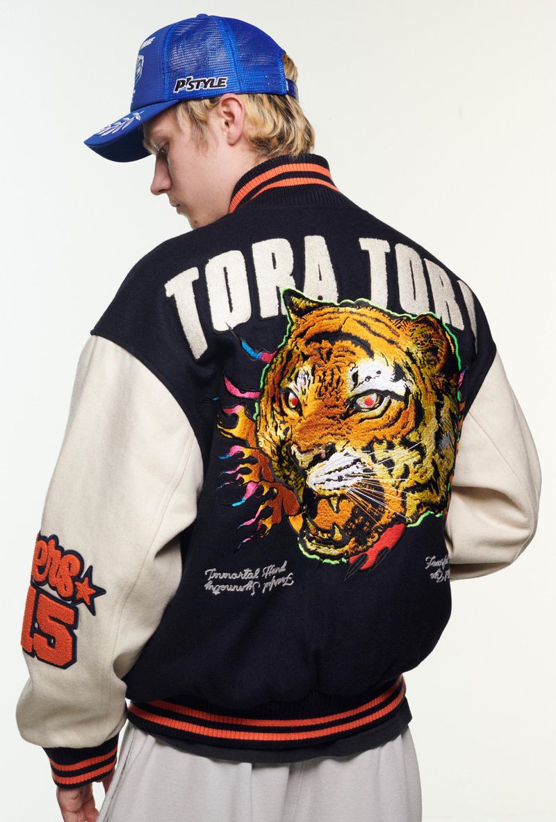 Man with blond hair wearing a blue baseball cap and black and white jacket with a tiger on the back, posing in a bright photo studio.