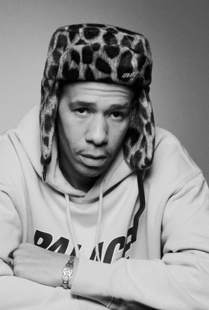 Monochrome portrait of a man wearing patterned hat and sweatshirt, arms crossed, against a grey backdrop, jewelry visible on wrist.