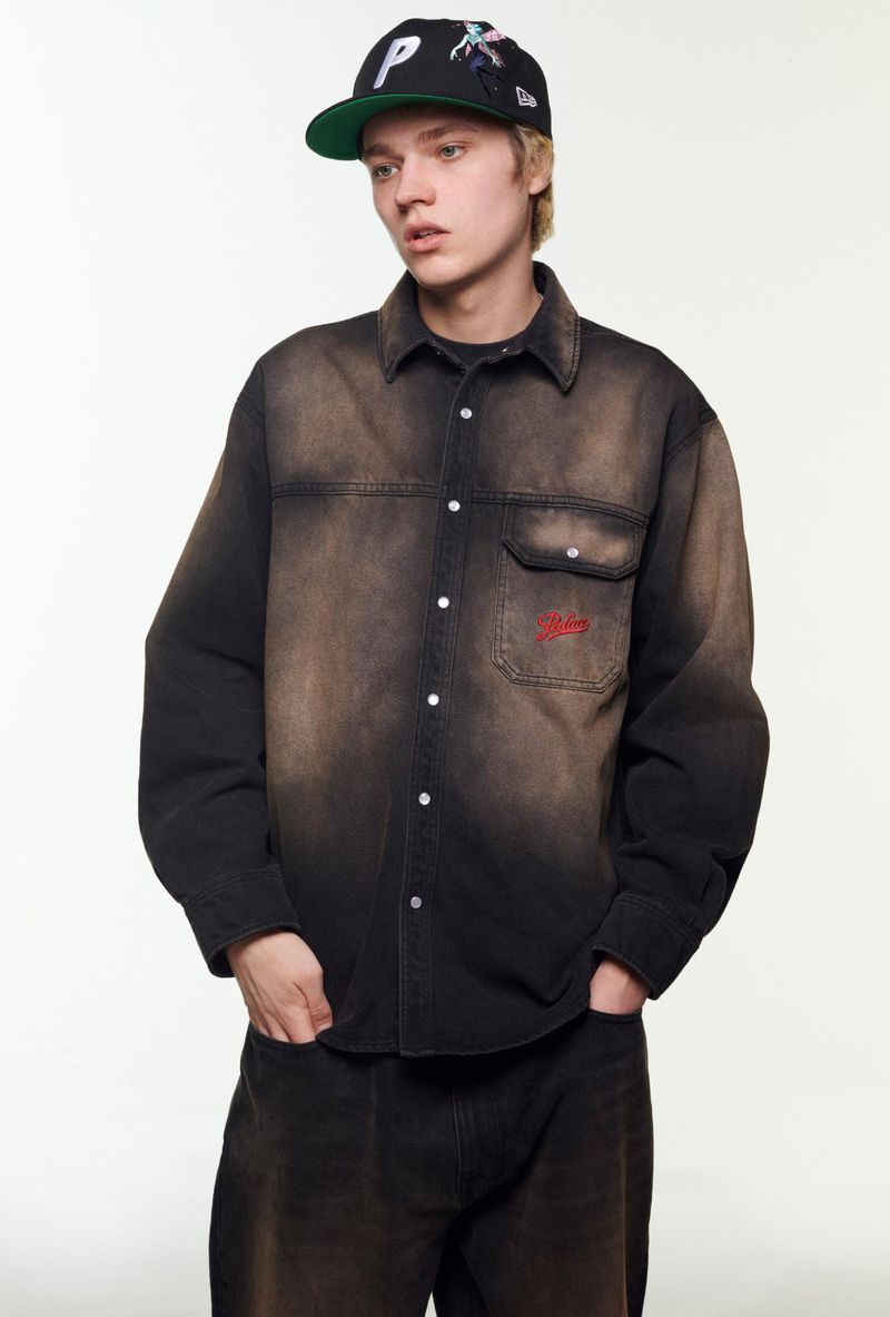 Man wearing a Palace baseball cap and faded button-down shirt poses indoors against a neutral background for a fashion shot.