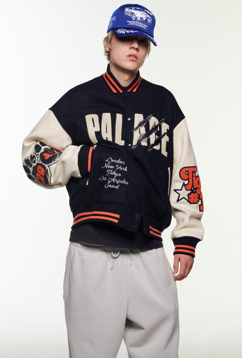 Young adult with blond hair wearing a baseball cap and Palace jacket with sweatpants in a studio with a white background.