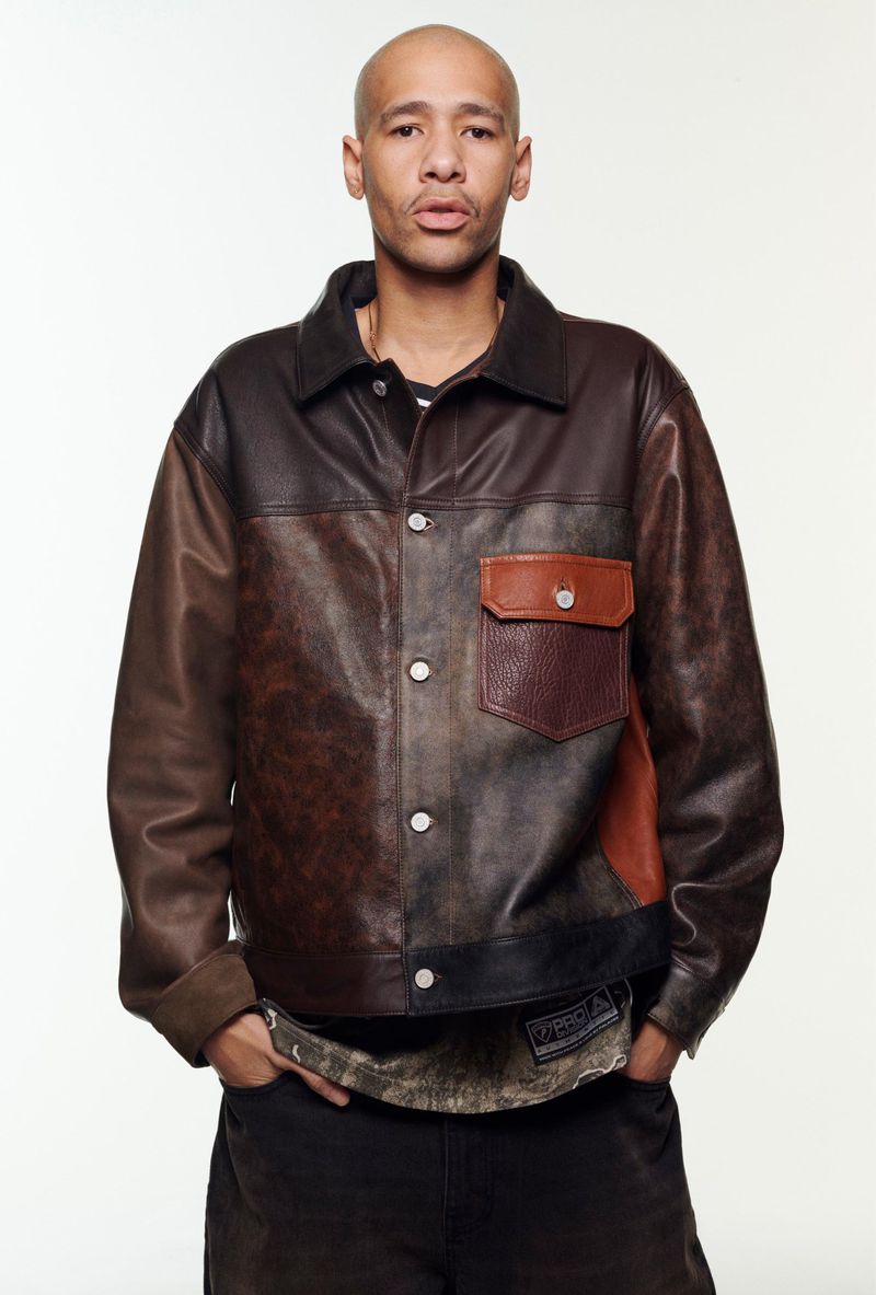 Adult male in a distressed brown leather jacket and black pants poses in front of a white backdrop in a studio setting.