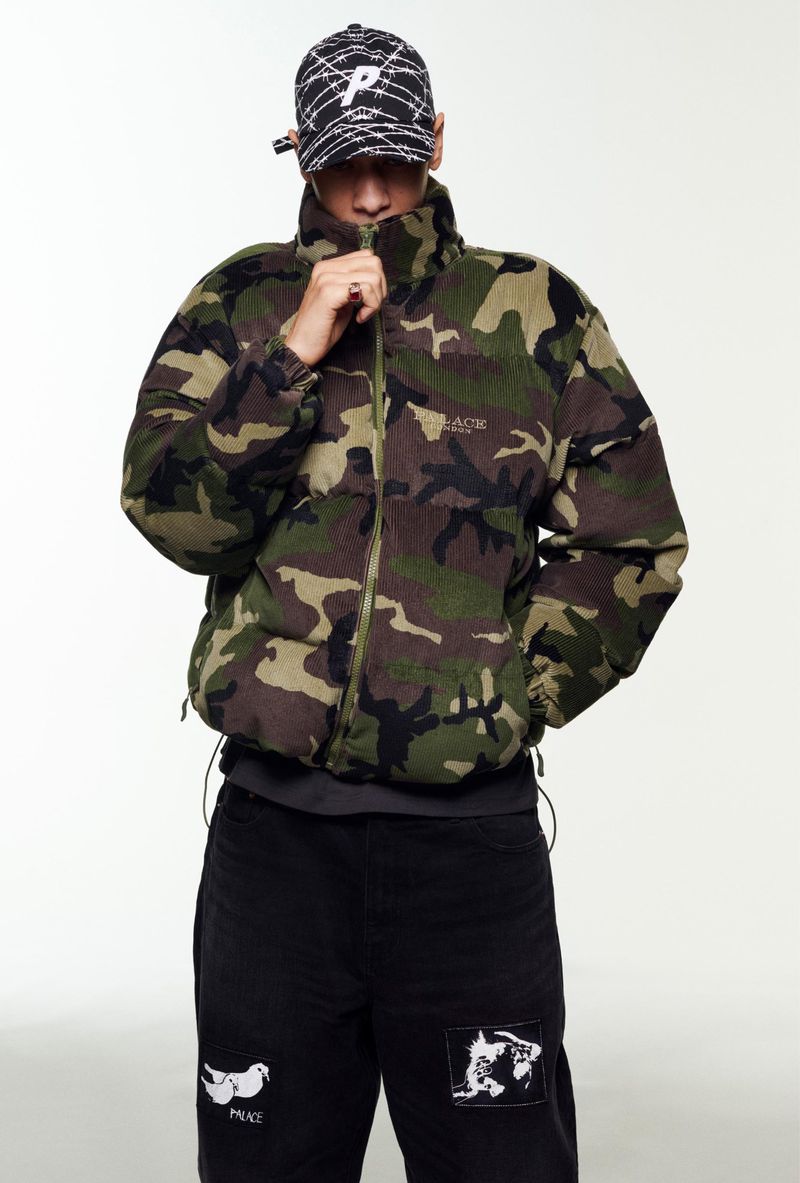 Adult male in a camouflage puffer jacket and patterned cap stands in a bright photo studio, pulling the jacket collar up to his face.