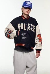 Model wearing a Palace varsity jacket and blue cap against a white background