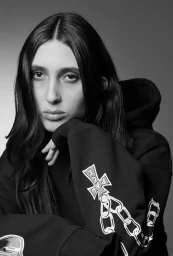 Portrait of a woman with long dark hair wearing a black hoodie with embroidered graphics against a grey background