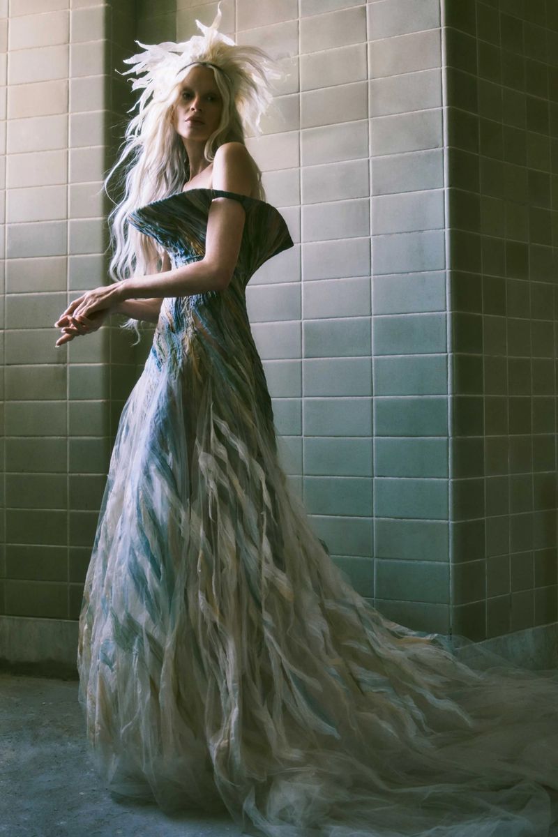 Woman in an elaborate off-the-shoulder gown with a feathered headpiece posing against a tiled wall in a fashion editorial style.