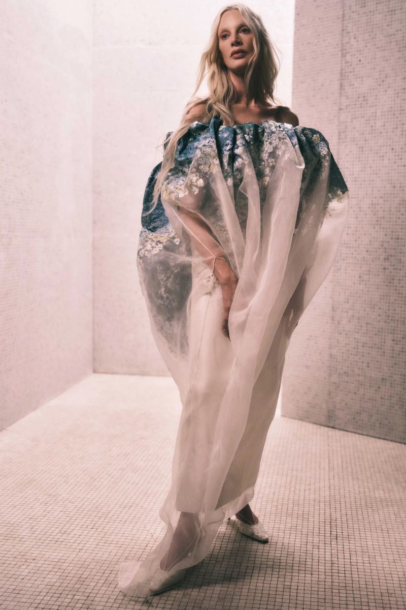 Woman in voluminous blue and white floral-patterned dress with sheer overlay, posing in an indoor tiled room.