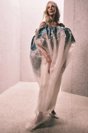 Woman in voluminous blue and white floral-patterned dress with sheer overlay, posing in an indoor tiled room.