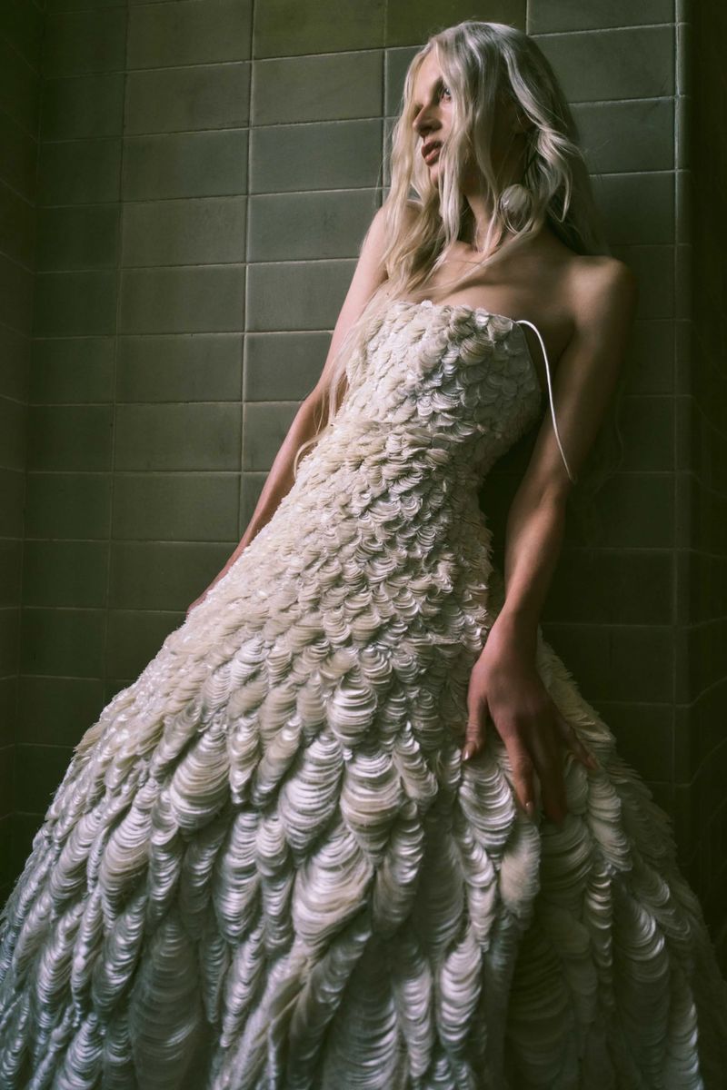 A fair-skinned woman with long blonde hair wears a formal strapless gown with shell-like details, leaning against a tiled wall indoors.