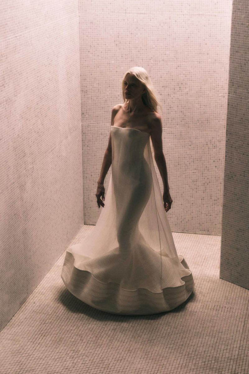 Elegant woman in a white gown stands in a tiled room. The soft lighting highlights the textures of the gown and the walls.