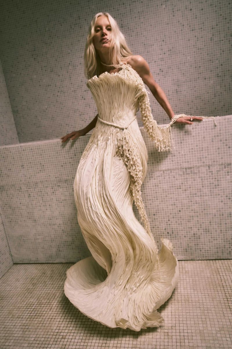 Blonde woman in full-length gown with one long sleeve, posing against tiled wall and floor, fashion shoot, beauty shot.
