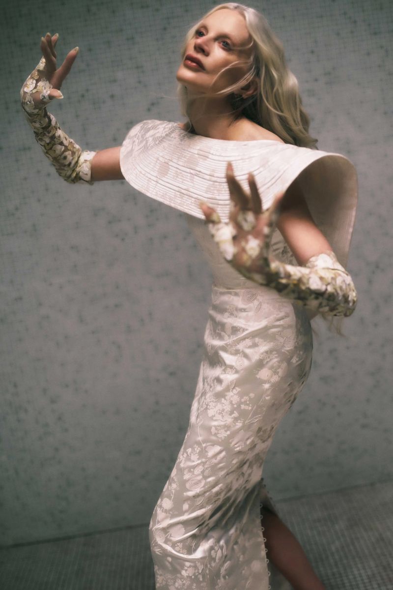 A woman with long, blonde hair wearing an elegant white dress and floral gloves stands against a mottled grey backdrop.