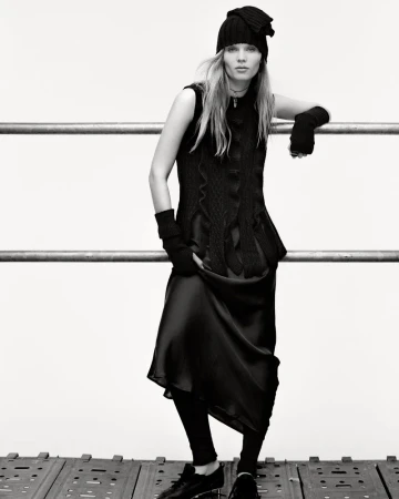 A solitary adult female in a black knit hat and sleeveless dress leans against a metal rail in a studio, casting a somber yet stylish gaze.