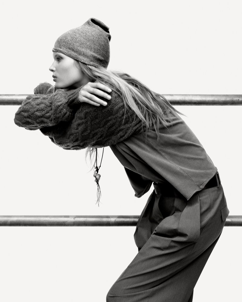 Monochrome portrait of a woman in a knit hat and sweater leaning on a metal bar, looking left with a neutral expression.