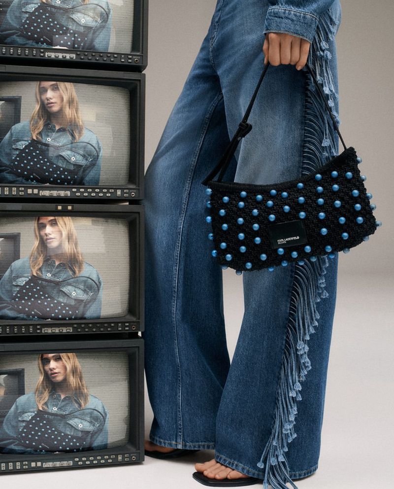 Model displays a black, beaded Karl Lagerfeld purse, standing beside stacked TVs showing the same image, wearing fringed-denim jeans.