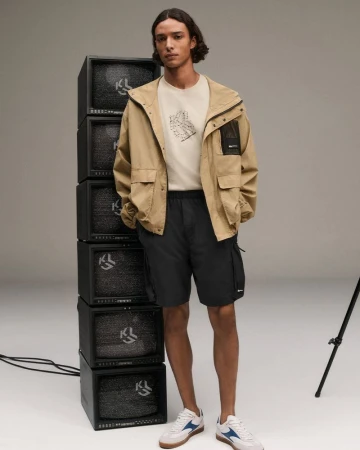 Male model poses in a tan jacket and black shorts next to a stack of old televisions in a studio with neutral grey background.