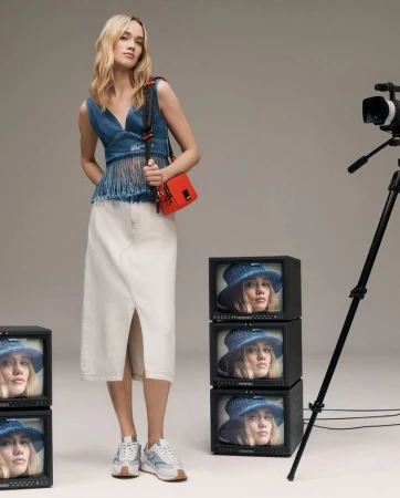 Blonde woman in denim top, long white skirt, sneakers stands in photo studio with video camera and monitors displaying her image.