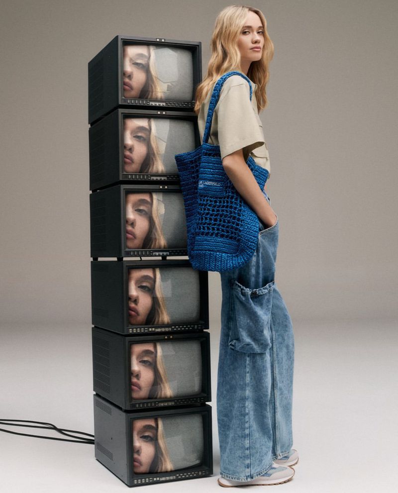 Young woman with a blue crocheted bag stands next to a stack of vintage televisions displaying her reflection in a neutral studio setting.