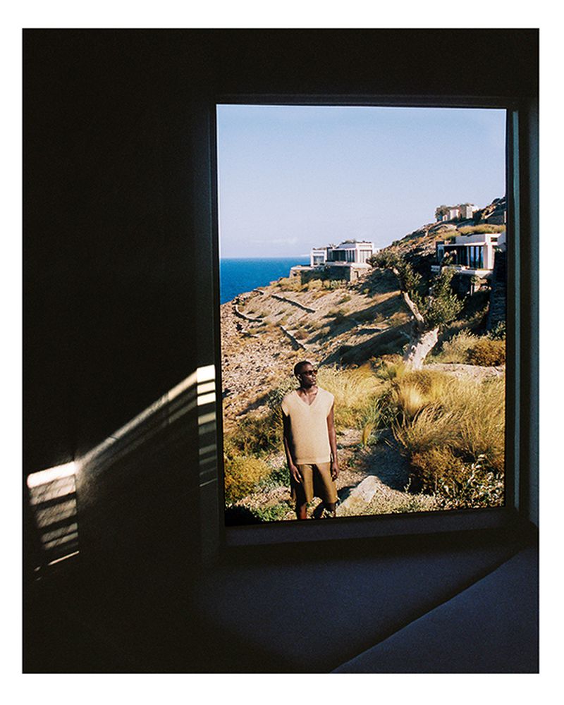 A man stands in a window with a view of hillside villas and the sea in a bright sunny landscape scene, featuring architectural and natural elements.