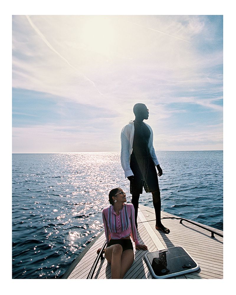 Two people on a yacht: An adult male stands while an adult female sits on the deck against a backdrop of ocean and blue sky.