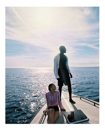 Two people on a yacht: An adult male stands while an adult female sits on the deck against a backdrop of ocean and blue sky.