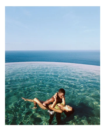 A biracial couple embraces in a turquoise infinity pool under a clear sky, overlooking the ocean, capturing a serene, romantic moment.