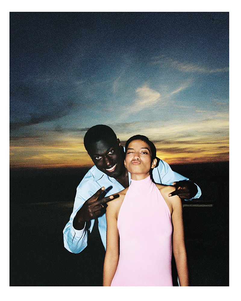 Smiling couple poses outdoors at sunset, with a man making a peace sign. The woman winks at the camera in a pink sleeveless dress.