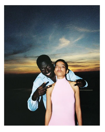 Smiling couple poses outdoors at sunset, with a man making a peace sign. The woman winks at the camera in a pink sleeveless dress.