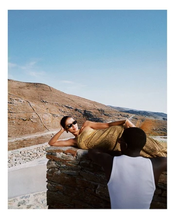 Woman in a gold dress poses on a stone wall in a sunny outdoor setting with a man in the foreground and a hillside backdrop.