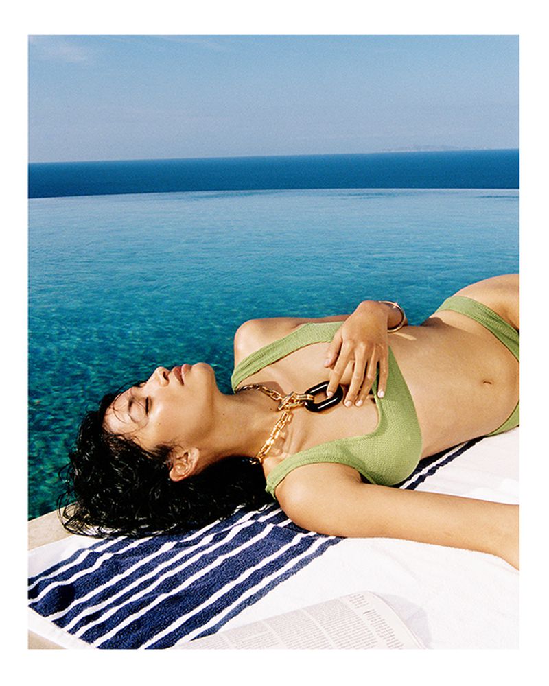 Woman in green bikini and gold necklace lounges on a blue-and-white striped towel by an infinity pool, with the ocean and sky in the distance.