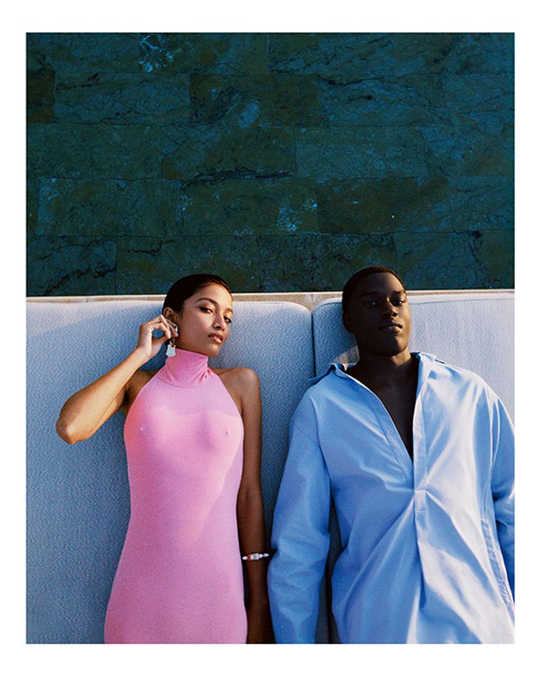 An adult interracial couple lounging together on a sofa with a blue textured wall behind them, posing in pink and blue attire in an outdoor setting.