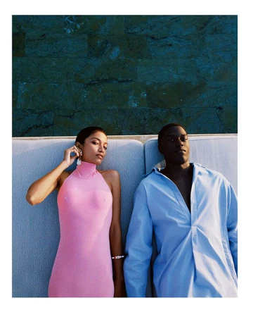 An adult interracial couple lounging together on a sofa with a blue textured wall behind them, posing in pink and blue attire in an outdoor setting.