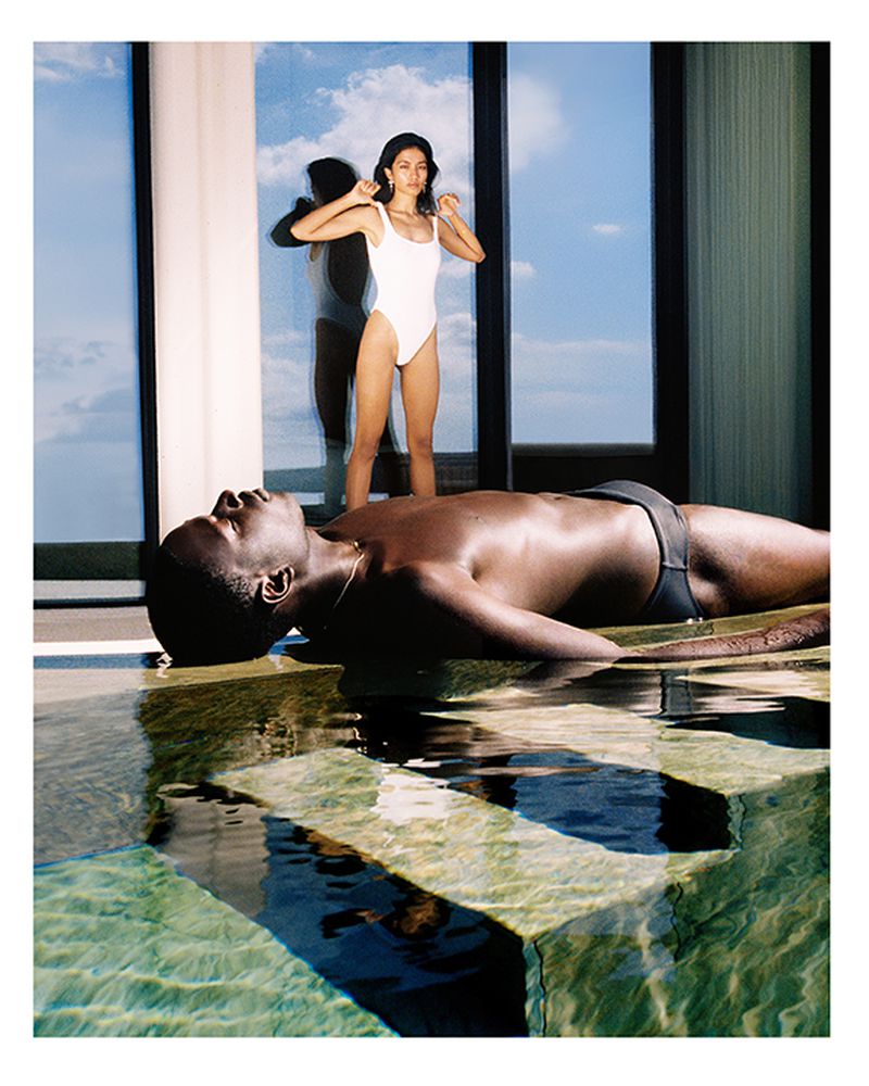 A dark-skinned man lies in shallow water near a pool's edge while a woman in a white swimsuit stands by a window with a sky view.
