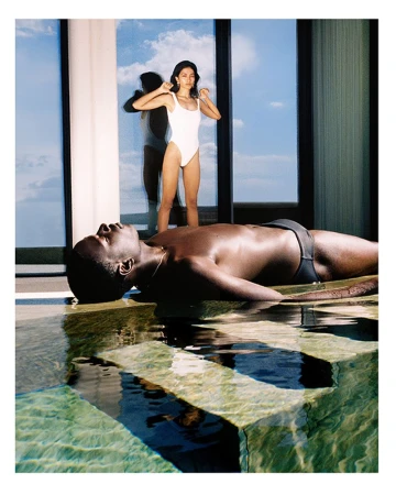 A dark-skinned man lies in shallow water near a pool's edge while a woman in a white swimsuit stands by a window with a sky view.