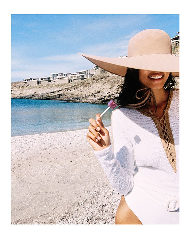 Woman in a beige sun hat enjoys a lollipop at the beach, posing in a white swimsuit with a knitted top under a clear blue sky.