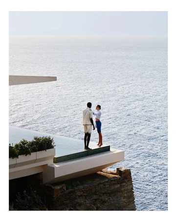 Woman adjusting man's collar on modern terrace with ocean view, bright daylight, elegant clothing, and minimalist design.