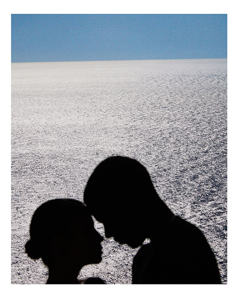 A silhouette of a couple standing close, their faces near each other, with the sea and sky in the background, and soft, diffused light.