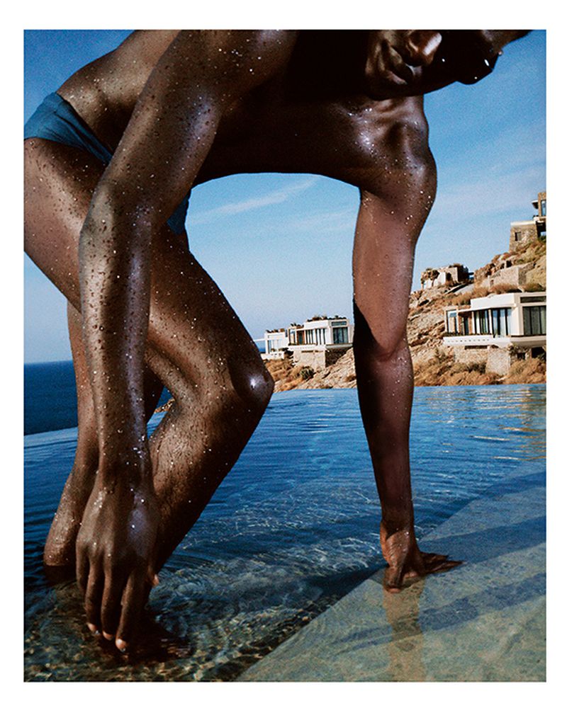 Close-up of a dark-skinned adult human entering an infinity pool outdoors with buildings and a blue sky in the background.
