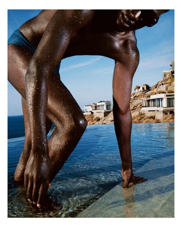 Close-up of a dark-skinned adult human entering an infinity pool outdoors with buildings and a blue sky in the background.