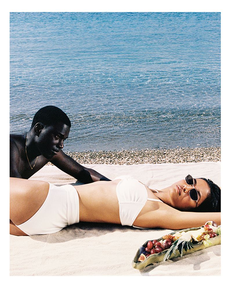 Two people relax on a beach near the ocean. An adult female in a swimsuit lies in the sun while an adult male sits beside her.