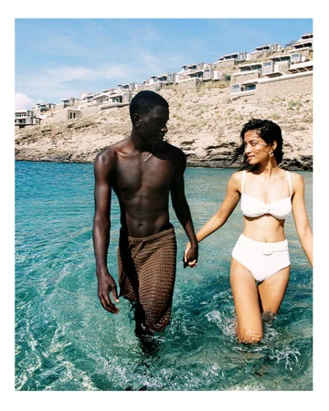 A couple walks hand-in-hand through shallow turquoise water, near a shore with bright white buildings on a rocky hillside under a clear sky.