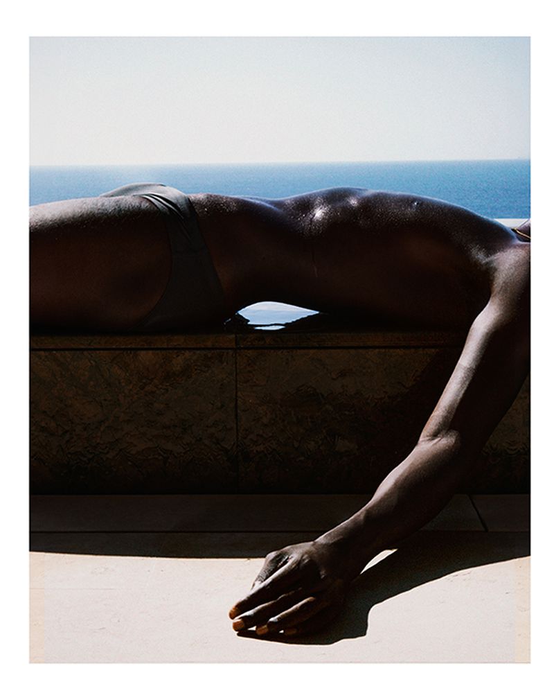 Torso of a person with dark skin lying on stone surface outdoors against a blurred background of blue sea and clear sky on a sunny day.
