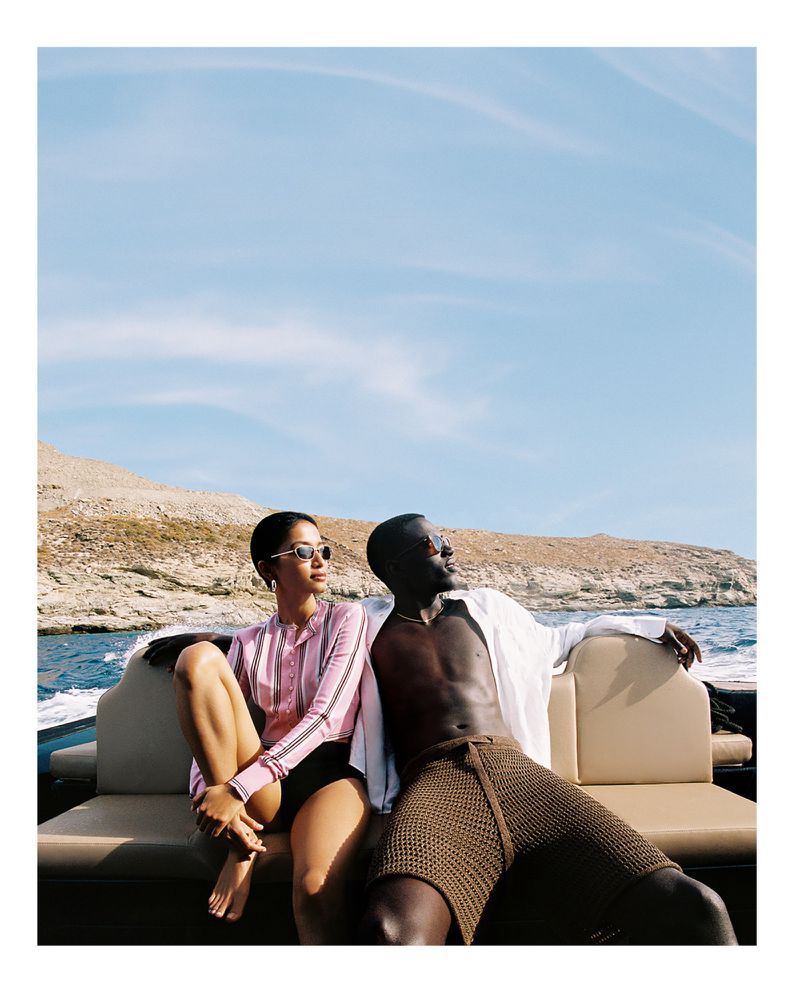 A couple sits on a boat with a rocky shoreline visible in the background. The woman wears sunglasses and a pink shirt, the man wears a white shirt.